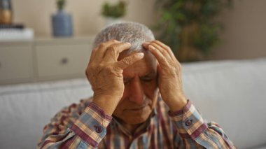 Ekose gömlekli, gri saçlı İspanyol bir adam oturma odasında oturmuş, elleri alnında sıkıntılı bir yüz ifadesi sergiliyor, kapalı bir arka planda bitkiler ve mobilyalarla oturuyor..