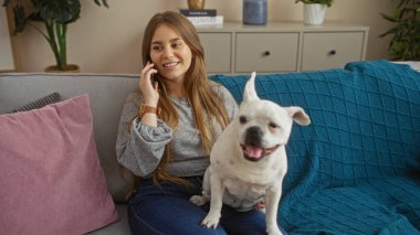 Sarışın, güzel bir kadın evindeki oturma odasındaki kanepede oturuyor ve yanında köpeğiyle telefonda konuşuyor..