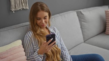 Rahat ev ortamında akıllı telefon kullanan genç bir kadın rahat modern iç mimariye sahip.