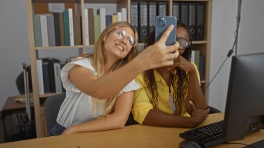 Kadınlar ofiste otururken, etrafında dosyalar ve bilgisayarla selfie çekiyorlar.