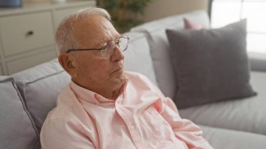 Olgun, gri saçlı bir adam rahat bir oturma odasında oturur, evinde huzurlu ve içsel bir anı somutlaştırır..