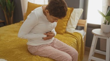 Kısa saçlı yaşlı bir İspanyol kadın yatak odasındaki bir yatağa oturur. Rahatsızlık içinde karnını tutar. Etrafı yumuşak mobilyalarla çevrilidir..