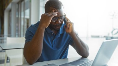 Ofiste çalışan genç bir adam, dizüstü bilgisayar kullanırken telefonla konuşuyor, modern çalışma alanında düşünceli bir ifade sergiliyor..