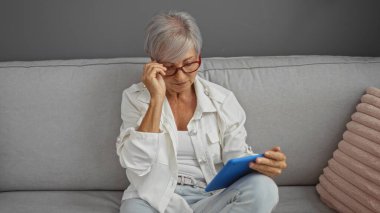 Kısa gri saçlı, gözlüklü ve beyaz gömlekli yaşlı bir kadın oturma odasındaki kanepede oturup tablet okuyor.