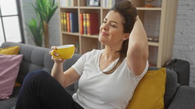 Rahat bir İspanyol kadın şık bir apartman dairesinde rahat bir kanepede bir fincan kahvenin tadını çıkarıyor.
