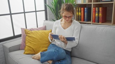 Rahat giyimli genç bir kadın rahat bir kanepede kitap okuyor, etrafı yastıklarla çevrili, iyi aydınlatılmış bir oturma odasında..