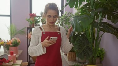 Kırmızı önlüklü genç bir kadın kapalı bir çiçekçinin yemyeşil bahçesinde akıllı telefon kullanıyor..