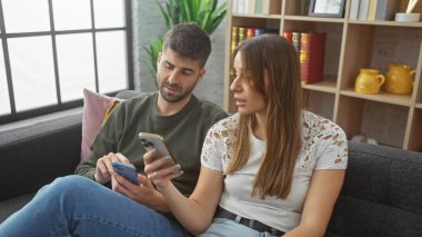 Bir çift evlerindeki bir kanepede dinleniyor, akıllı telefonlar ile meşgul, modern kapalı yaşam tarzını tasvir ediyor..