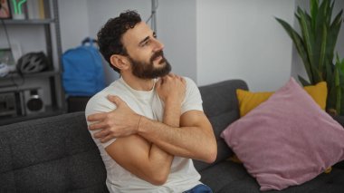 Düşünceli sakallı bir adam modern bir oturma odasında bir bitkiyle rahatça oturuyor.