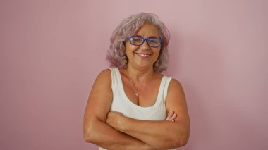 Gri saçlı ve gözlüklü olgun İspanyol bir kadın, kolları kapalı pembe bir arka plana yaslanmış güvenle ayakta duruyor..