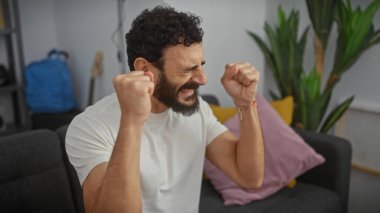 Heyecanlı sakallı adam modern bir oturma odasında başarıyı kutluyor.