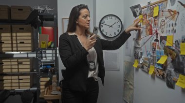 Hispanic woman detective analyzing evidence on board in a police department office. clipart