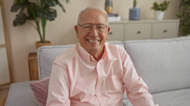 Gri saçlı, orta yaşlı adam sıcak bir şekilde gülümsüyor, rahat bir oturma odasında oturuyor..