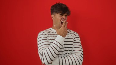 Yakışıklı, genç, İspanyol bir adam koyu kırmızı bir duvara yaslanmış, çizgili bir gömlek giymiş, yorgun bir ifadeyle izole edilmiş..