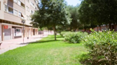 Bulanık açık hava sahnesi. Güneşli bir şehir ortamında çözünmüş binalar, ağaçlar, çalılar ve çimenler.