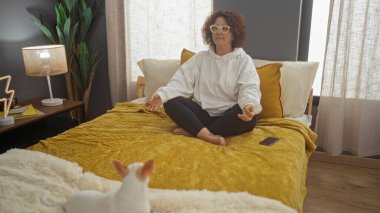 Orta yaşlı bir kadın yatakta bir chihuahua köpeğiyle meditasyon yapıyor. Etrafı kapalı bitkilerle ve yumuşak mobilyalarla çevrili. Huzurlu bir ev dekorasyonu yaratıyor..