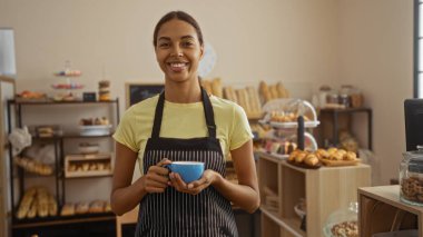 Taze ekmek ve hamur işleriyle dolu bir pastanede elinde fincanla gülümseyen önlüklü genç kadın.