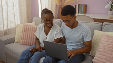 Anne ve oğlu rahat bir oturma odası kanepesinde oturuyor, gülümsüyor ve dizüstü bilgisayar kullanıyor, Afro-Amerikan aile sevgisinin altını çiziyor ve içeride kaynaşıyor..