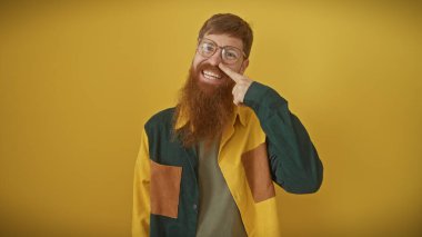 Yakışıklı kızıl sakallı adam sarı arka planda kendine güvenerek poz veriyor, parmağıyla burnuna dokunuyor..