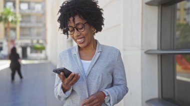 Kıvırcık saçlı ve gözlüklü güzel bir Afro-Amerikalı kadın modern binaların bulunduğu bir şehirde şehir sokağında dururken telefonunu kontrol ediyor..