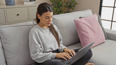 Genç bir İspanyol kadın oturma odasında kanepede dizüstü bilgisayar kullanarak oturuyor. Ev ortamında, rahat bir atmosferle yakalanmış..