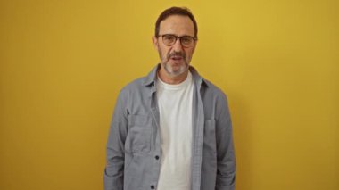 İspanyol orta yaşlı gözlüklü bir adam ayakta dikiliyor ve şaşırmış görünüyor. Sarı arka plan, şaşırmış yüz ifadesi, işaret eden mutlu ruh hali.