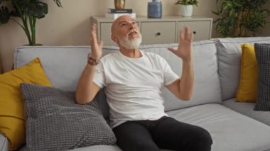 Gri saçlı ve sakallı bir adam, bitki ve yastıklarla çevrili, modern bir iç mekanı olan bir salonda otururken hayal kırıklığına uğramış görünüyor..