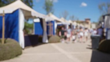 Mallorca, İspanya 'da mavi çadırları olan güneşli bir açık hava pazarının keyfini çıkaran insanlar, canlı atmosferi ve toplumsal bağlılığı vurgulamak için bulanık ve odaksız unsurlar sunuyorlar.