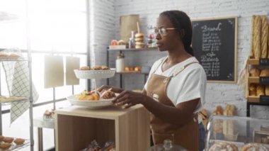 Genç Afrikalı Amerikalı kadın gözlüklü ve önlüklü. Kapalı mekanlarda ekmek ve hamur işi sergilenen rahat bir pastanede hamur işi düzenliyor.