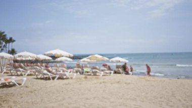 Güneşli bir günde arka planda kumlu kıyıları ve sakin okyanus dalgalarıyla şemsiyelerin altında dinlenen insanların olduğu bulanık plaj sahnesi