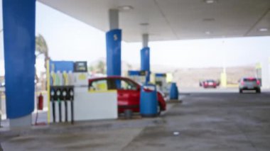 Benzin istasyonunda, gölgeliğin altında kırmızı bir araba ve arka planda diğer araçlar park halindeyken görüntüsü bulanık.