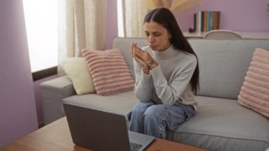 Oturma odasındaki kanepede oturan genç bir kadın, dizüstü bilgisayara odaklanmış, yakınlardaki bir pencereden içeri doğal ışık süzülüyor, arka planda yumuşak yastıklar ve bir kitaplık ile çevrili.