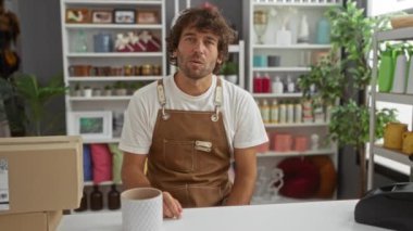 Genç adam bir ev dekorasyonu mağazasında çalışıyor, önlük giyiyor ve raflarda çeşitli dekoratif eşyalarla çevrili beyaz bir seramik vazo tutuyor.