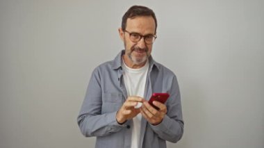 İspanyol bir adam, orta yaşlı, akıllı telefon tutuyor, işaret dilini elinde tutuyor, mutlu yüz ifadesi, parmaklarından bakıyor, gülümsüyor, beyaz arka planda soyutlanıyor.