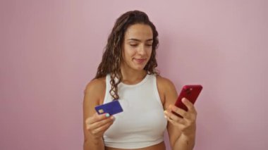 Elinde cep telefonu ve kredi kartı olan genç bir kadın izole edilmiş pembe bir arka plana gülümsüyor.