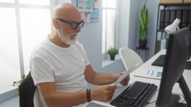 Ofisteki olgun gri saçlı adam, arka planda bilgisayar ve çalışma masası olan bir belgeye bakarken gözlüklerini ayarlıyor..
