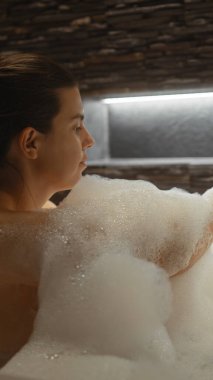 İçeride köpüklü bir banyoda rahatlayan, dinginlik ve özgüven saçan genç bir kadının yan profili..