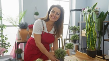 Kırmızı önlüklü, gülümseyen bir İspanyol kadın ev ortamında yemyeşil çiçeklerle çevrili, kapalı bir yerde bitkiler düzenler..