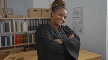 Ofiste güvenle gülümseyen, geleneksel yargıç cübbesi giyen, kolları çapraz duran, kitaplıklar ve yasal belgelerle çevrili, profesyonel bir çevreyi tasvir eden bir kadın.