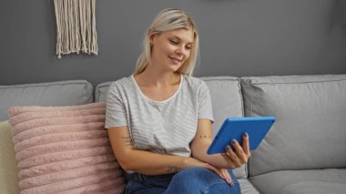 Oturma odasında bir kanepede oturan genç bir kadın, elinde mavi bir tablet, gülümsüyor, rahat bir dekor ile çevrili, sarı saçlı, çizgili bir gömlek ve bir ev ortamında günlük kot pantolon giyiyor.