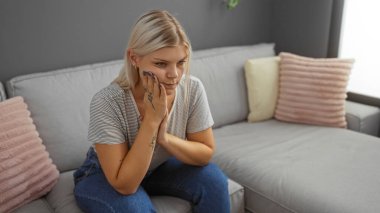 Genç bir kadın oturma odasında bir kanepede oturuyor, düşünceli ve düşünceli görünüyor, sarı saçlı sıradan kıyafetler giyiyor, modern bir apartman dairesi ve rahat bir ev dekorasyonu sunuyor.