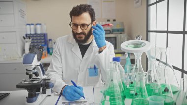 Laboratuvar önlüklü orta yaşlı sakallı bir adam, ekipmanlarla çevrili bir bilim laboratuarında notlar alıyor.