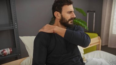 Sakallı bir adam modern bir yatak odası ortamında omuz ağrısı hisseder..