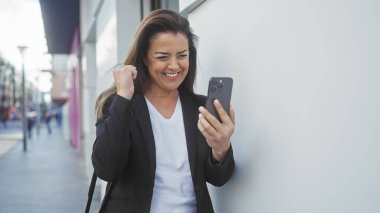 Coşkulu İspanyol bir kadın, bir şehir caddesinde akıllı bir telefonla başarıyı kutluyor.