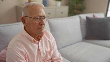 Oturma odasında oturan gri saçlı, gözlüklü ve pembe gömlekli, ev dekorunda dalgın görünen yaşlı beyaz bir adam..