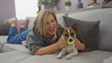 Sarışın, orta yaşlı bir kadın oturma odasındaki kanepede uzanır, rahat bir evde küçük köpeğiyle oynar..