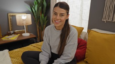 Çekici, esmer, genç bir kadın, rahat bir yatak odasında, rahat bir yatakta, kameraya sıcak bir şekilde gülümsüyor, etrafında peluş yastıklar ve sıcak ev dekorasyonu var..