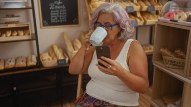 Woman drinking coffee while using her smartphone in a cozy bakery shop surrounded by fresh bread and pastries. clipart