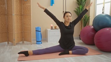 Spor salonunda yoga yapan genç Çinli bir kadın spor kıyafetlerini giyiyor ve spor aletleri ve dekorlarla çevrili bir paspasın üzerinde esneme hareketi yapıyor..