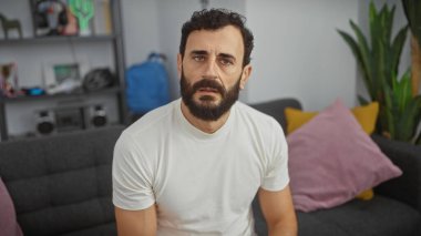 Modern bir oturma odasında otururken beyaz gömlekli, sakallı, orta yaşlı bir adam dalgın görünüyor..